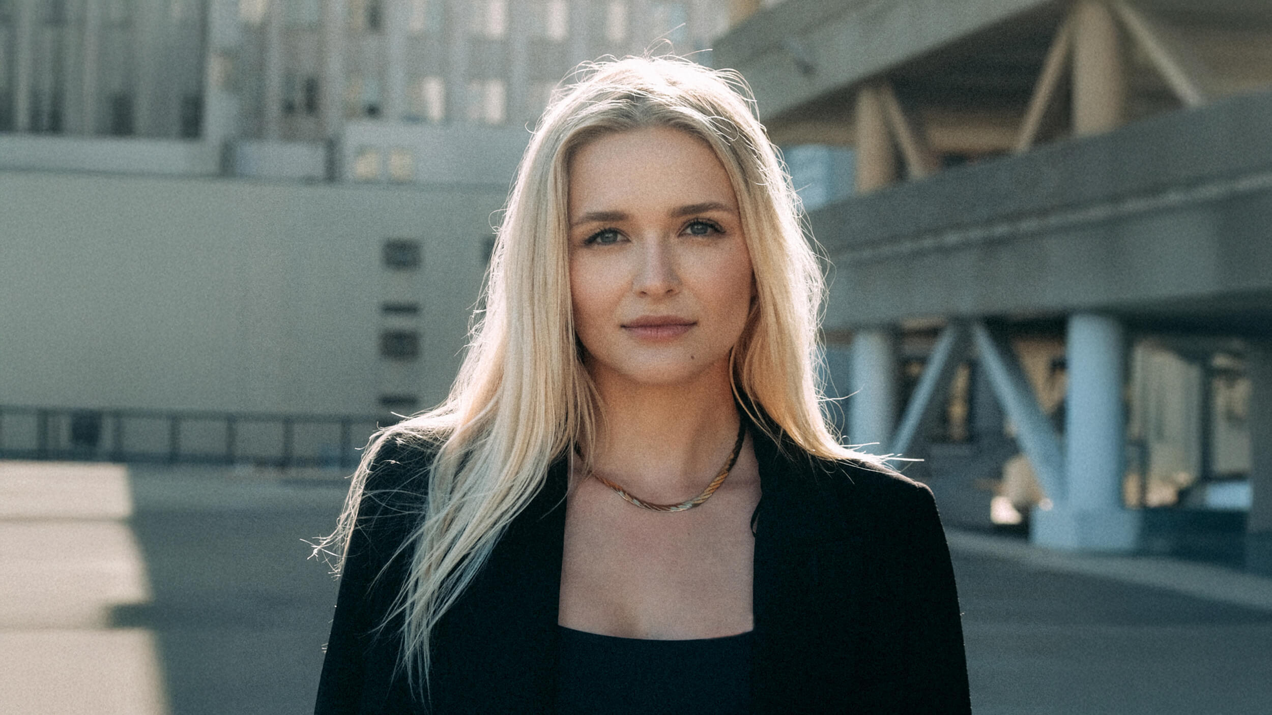 A woman with long blonde hair, wearing a black top and blazer, stands outdoors in front of modern buildings on a sunny day.