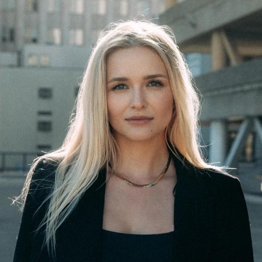 A woman with long blonde hair, wearing a black top and blazer, stands outdoors in front of modern buildings on a sunny day.