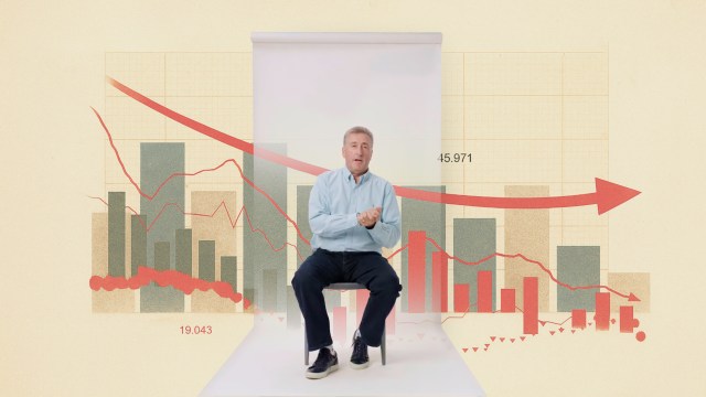 A man sits on a stool in front of a backdrop displaying a declining graph with bar charts and red trend lines, suggesting a downward trend in data.