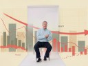 A man sits on a stool in front of a backdrop displaying a declining graph with bar charts and red trend lines, suggesting a downward trend in data.