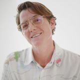 A person with short brown hair and glasses smiles at the camera, wearing a light-colored, patterned button-up shirt against a plain white background.