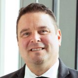 A man in a dark suit, white shirt, and black tie smiles while standing indoors near large windows.