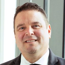 A man in a dark suit, white shirt, and black tie smiles while standing indoors near large windows.