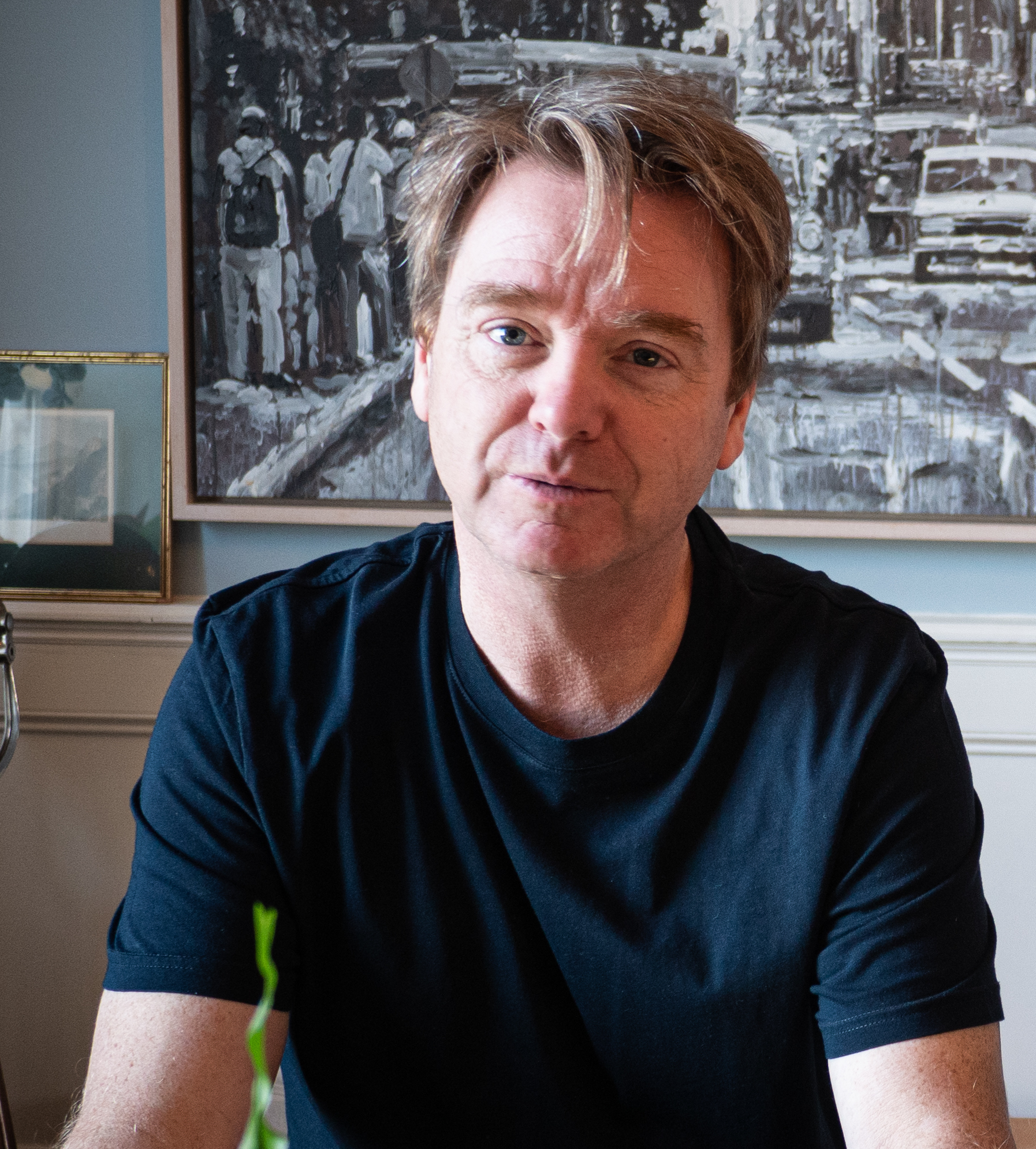 A man with light brown hair wearing a black t-shirt sits indoors in front of a black and white painting and a framed photo.