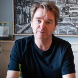 A man with light brown hair wearing a black t-shirt sits indoors in front of a black and white painting and a framed photo.