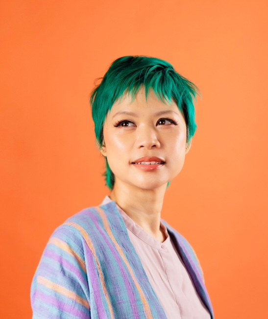 Person with short green hair and a pastel striped jacket looks slightly to the side against a solid orange background.