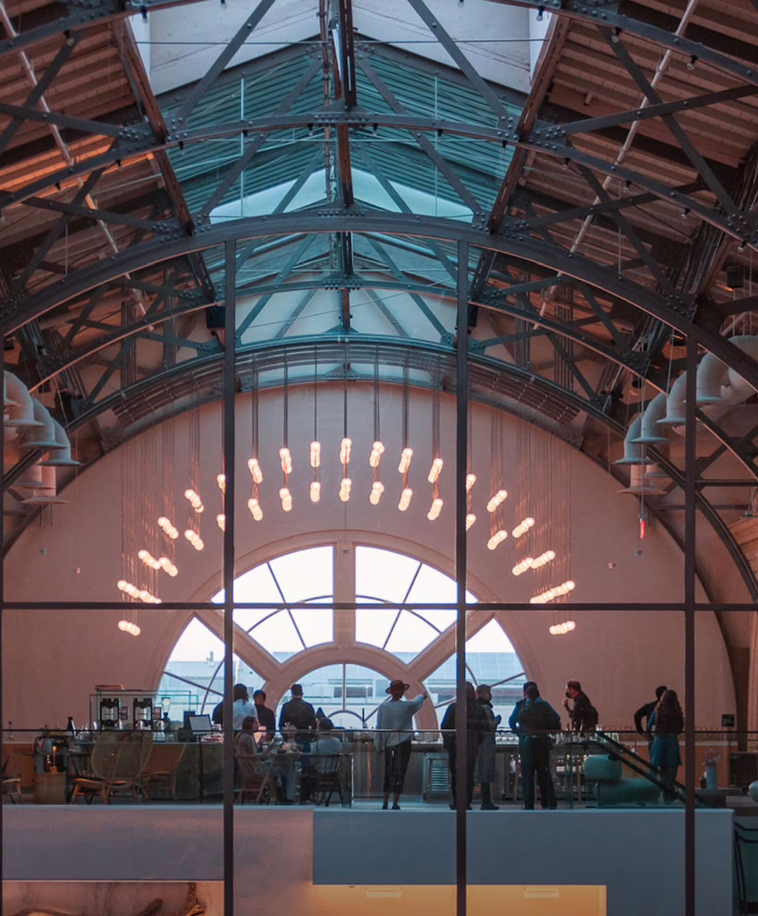 People standing and sitting inside a spacious building with a high arched ceiling, large windows, and hanging lights.