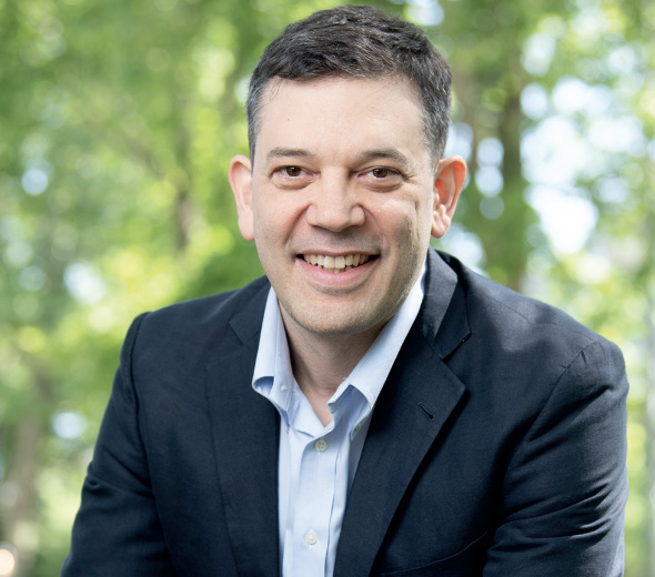 A man in a dark blazer and light blue shirt is smiling outdoors with green trees in the blurred background.