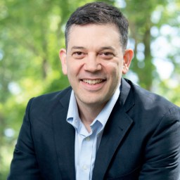 A man in a dark blazer and light blue shirt is smiling outdoors with green trees in the blurred background.