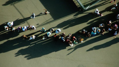 Aerial view of two groups of people waiting in line and walking on a paved surface, casting long shadows in the sunlight.