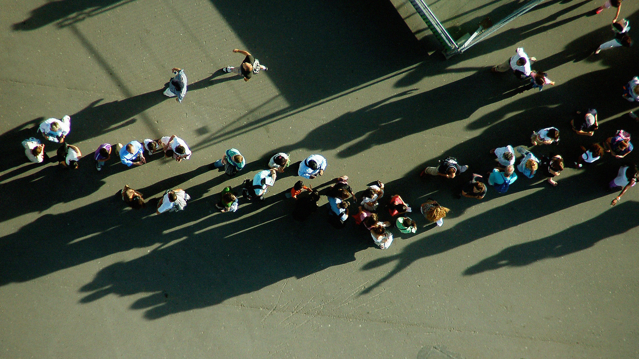 Aerial view of two groups of people waiting in line and walking on a paved surface, casting long shadows in the sunlight.