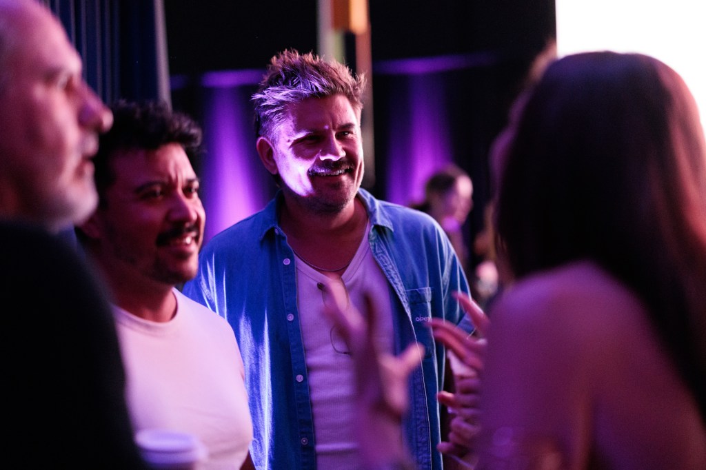 Three people stand talking in a dimly lit indoor setting with purple lighting, while a fourth person gestures in the foreground.