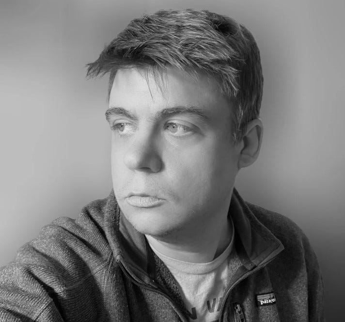 A man with short hair and a zip-up jacket looks to his left against a plain background in a black and white photo.