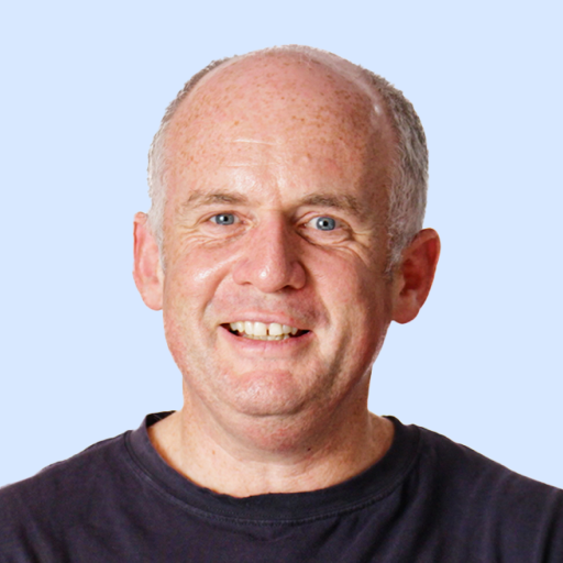 A middle-aged man with short gray hair and a receding hairline is smiling, wearing a dark t-shirt against a light blue background.