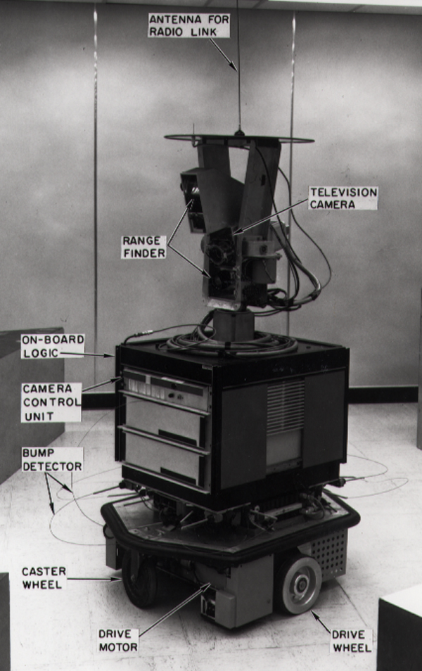 Black and white photo of an early mobile robot with labeled components including a television camera, antenna, range finder, drive wheels, control units, bump detector, and motors.