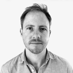 A man with short hair and light facial hair wears a light-colored button-up shirt and looks directly at the camera against a plain white background.