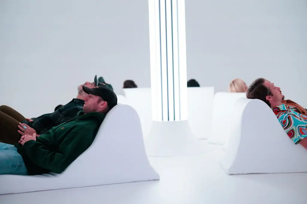 Several people recline on white lounge chairs in a bright, minimalist room with a tall, illuminated column in the center.