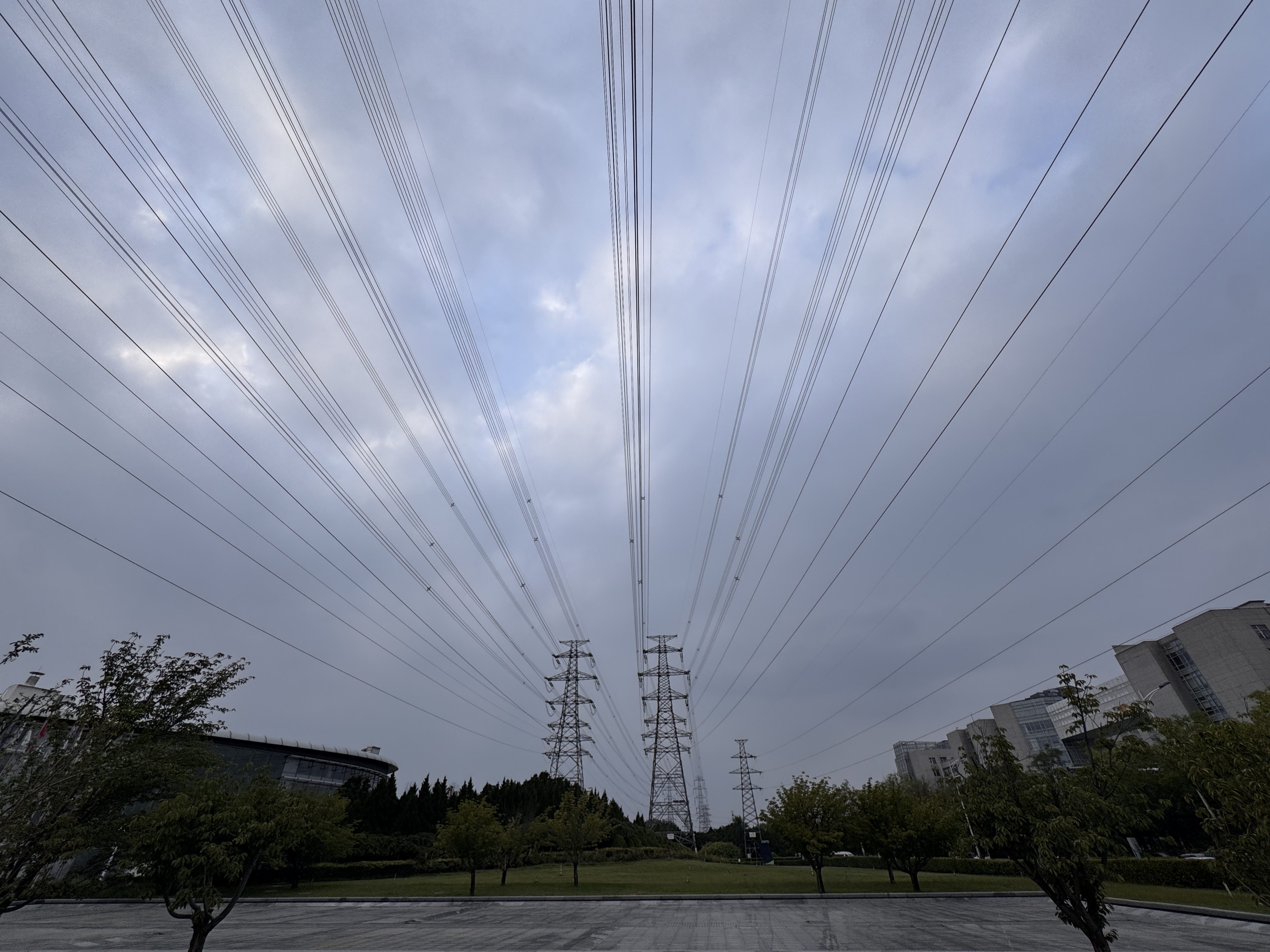 Multiple high-voltage power lines stretch across a cloudy sky above an urban area in China, with buildings, trees, and a wide paved surface below.