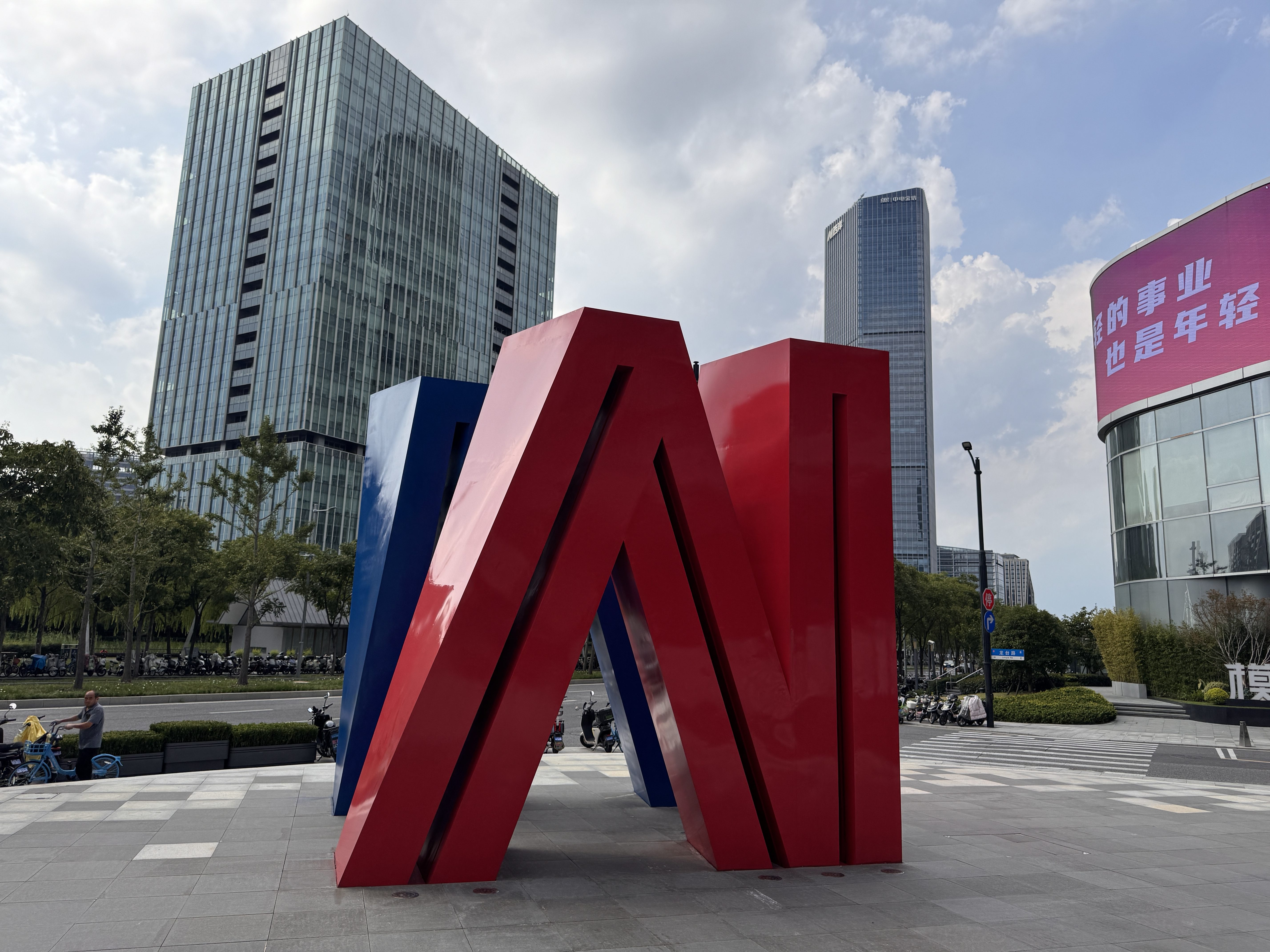 Large red and blue abstract metal sculpture in an urban plaza in China, with modern glass office buildings in the background under a partly cloudy sky.