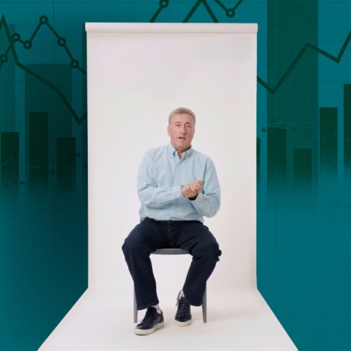 A man in a light blue shirt sits on a chair in a white studio with a teal background featuring graphs and charts.