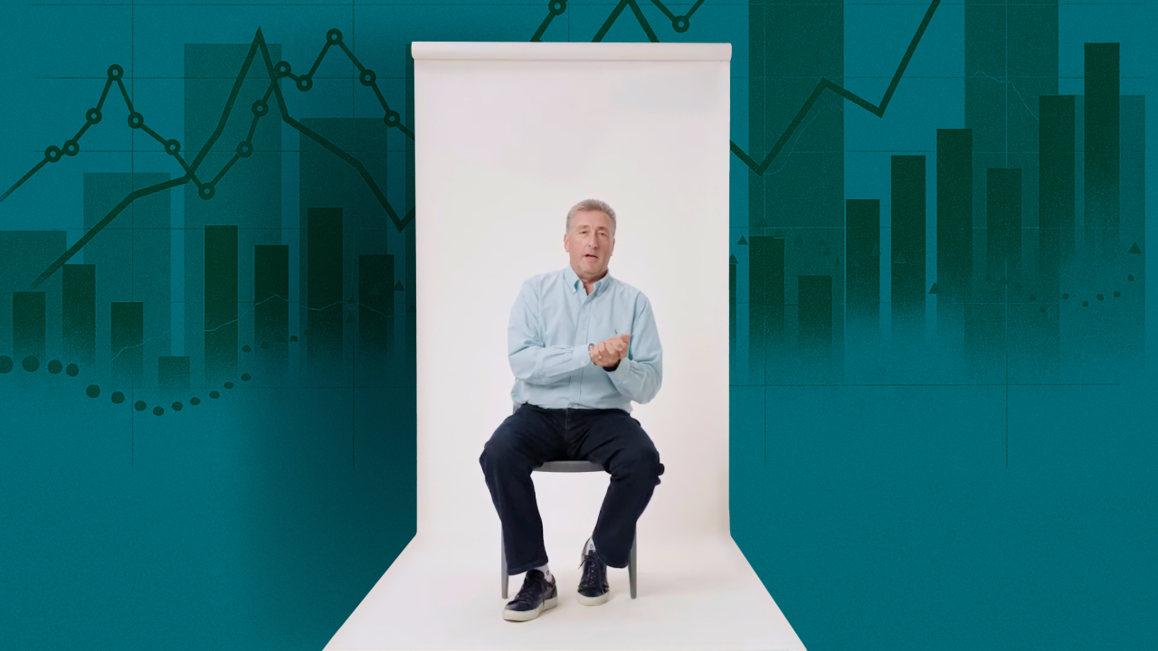 A man in a light blue shirt sits on a chair in a white studio with a teal background featuring graphs and charts.