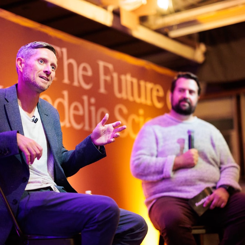 Two men sit on stage in discussion; one gestures while talking, the other holds a microphone. The backdrop reads “The Future of Psychedelic Sci.” Warm lighting illuminates the scene.