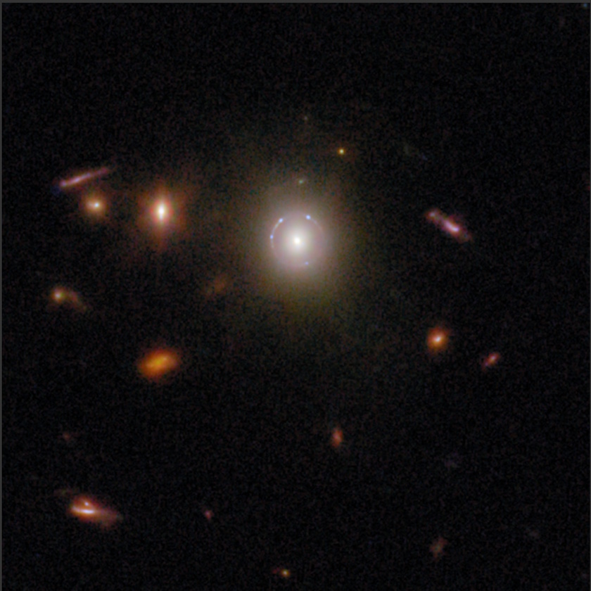 A galaxy cluster is shown in deep space, captured by COSMOS-Web JWST, with a bright central galaxy surrounded by smaller, reddish galaxies and faint circular gravitational lens distortions around the center.
