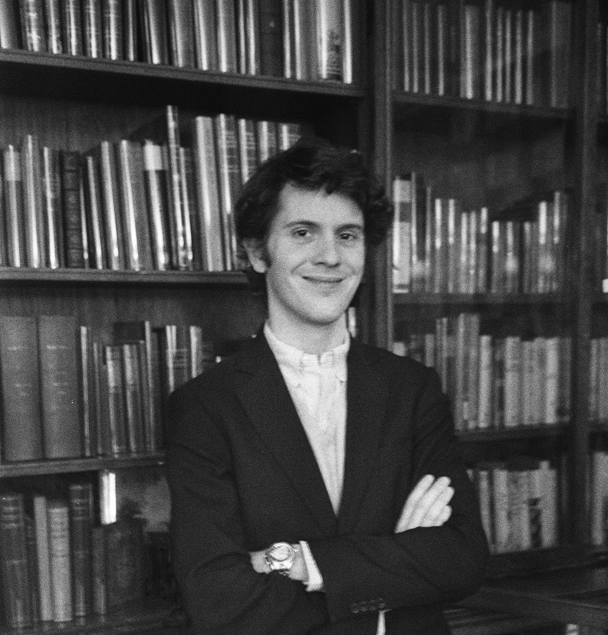 A young man in a suit jacket stands with arms crossed, smiling in front of bookshelves filled with books.