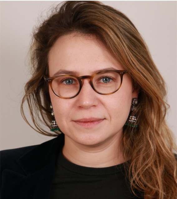 A woman with light brown hair, wearing round glasses, a dark top, and dangling earrings, looks directly at the camera against a neutral background.