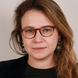 A woman with light brown hair, wearing round glasses, a dark top, and dangling earrings, looks directly at the camera against a neutral background.