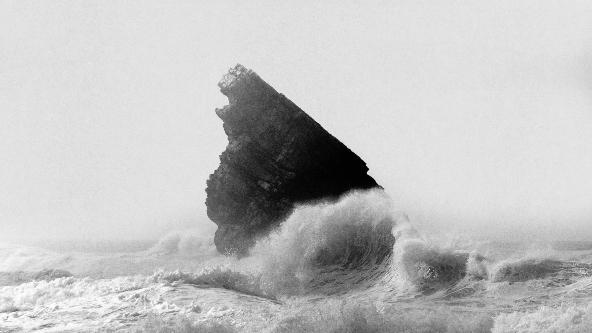 A large jagged rock juts out of rough ocean waves, with water crashing against its base under a cloudy sky—standing firm like true leadership amidst the storm.