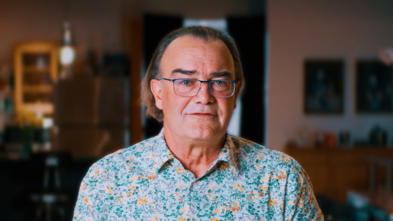 A middle-aged man with glasses and long hair stands indoors, wearing a floral-patterned shirt. The background is softly blurred with kitchen and living room elements visible.