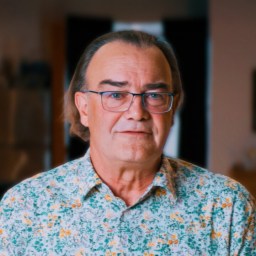 A middle-aged man with glasses and long hair stands indoors, wearing a floral-patterned shirt. The background is softly blurred with kitchen and living room elements visible.