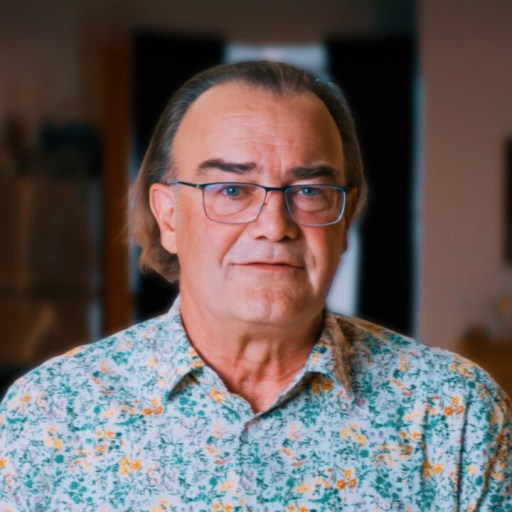 A middle-aged man with glasses and long hair stands indoors, wearing a floral-patterned shirt. The background is softly blurred with kitchen and living room elements visible.