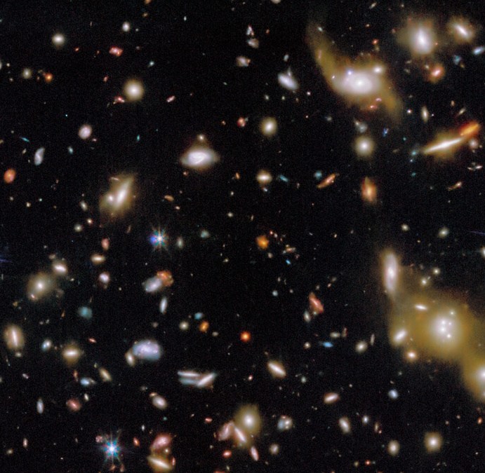 A view of deep space showing numerous galaxies of various shapes and sizes scattered across a dark background.