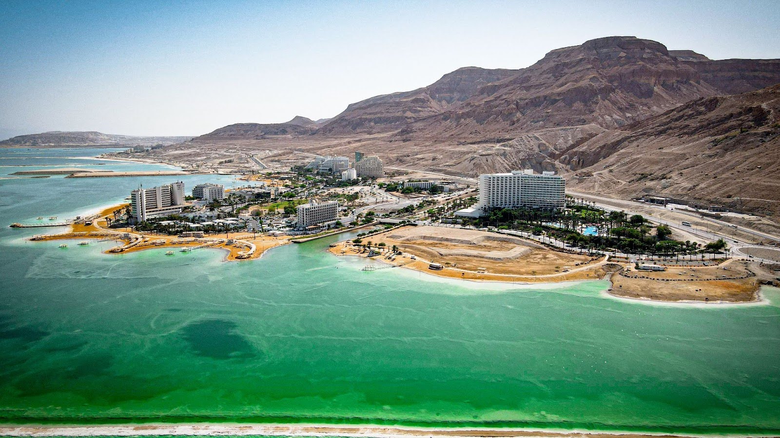 Aerial view of seaside hotels and resorts along a greenish-blue shoreline, bordered by arid mountains in the background.