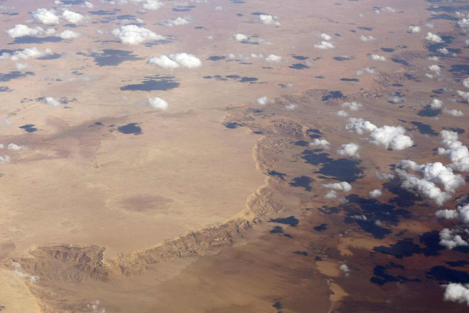 Aerial view of a vast desert landscape with scattered clouds casting shadows over rocky ridges and flat sandy terrain.