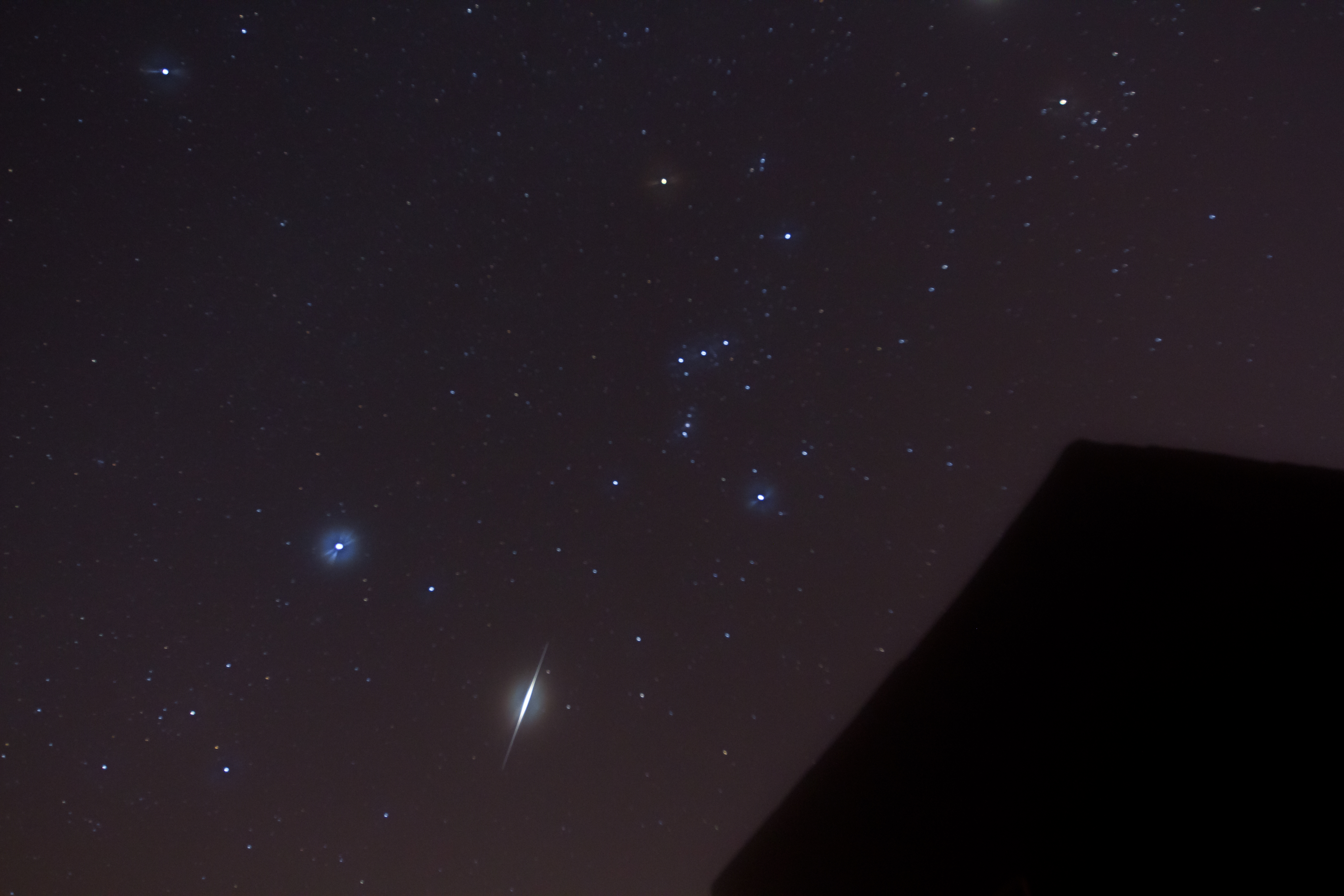 Night sky showing the Orion constellation, several bright stars, and a visible shooting star streak above a dark silhouette of a building, reminiscent of meteor showers from Halley's Comet.