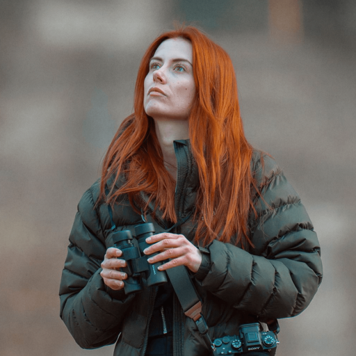 A woman with long red hair, wearing a puffer jacket, stands outdoors with passion in her gaze—holding binoculars and looking up as a camera hangs from her neck.