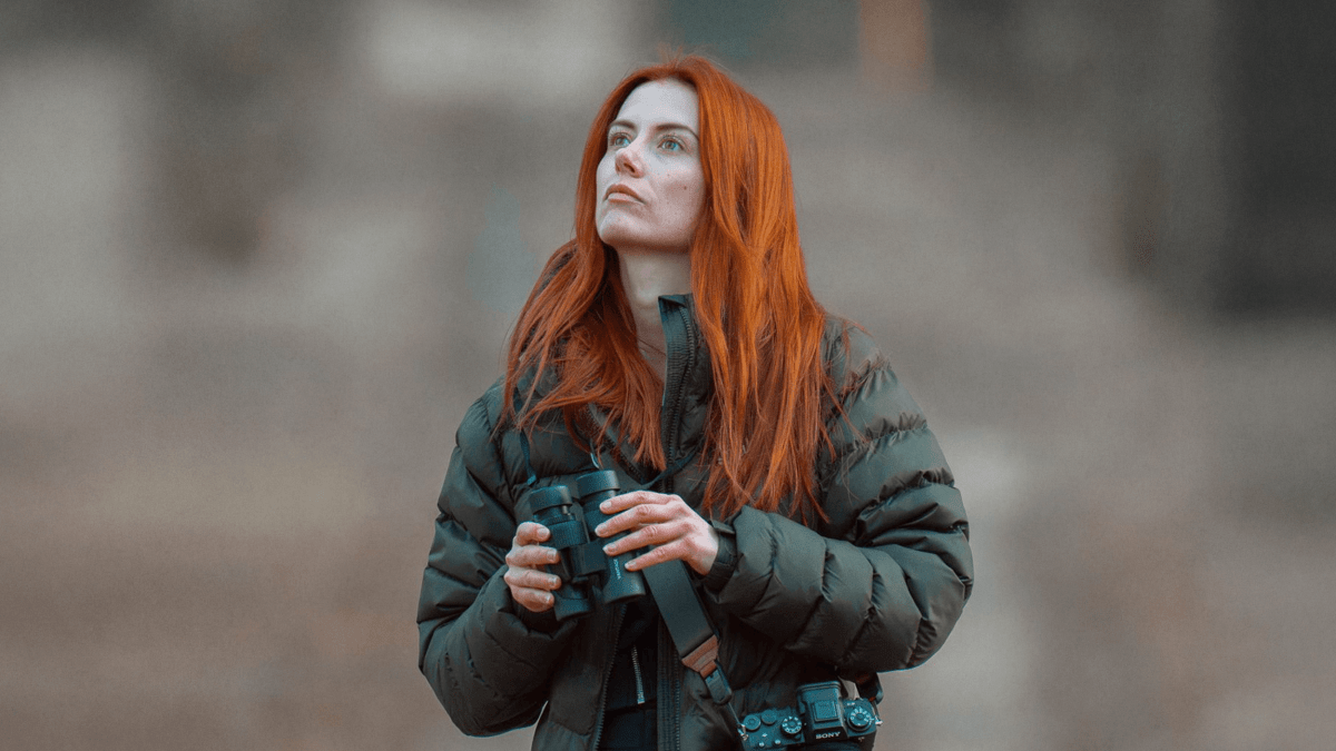 A woman with long red hair, wearing a puffer jacket, stands outdoors with passion in her gaze—holding binoculars and looking up as a camera hangs from her neck.