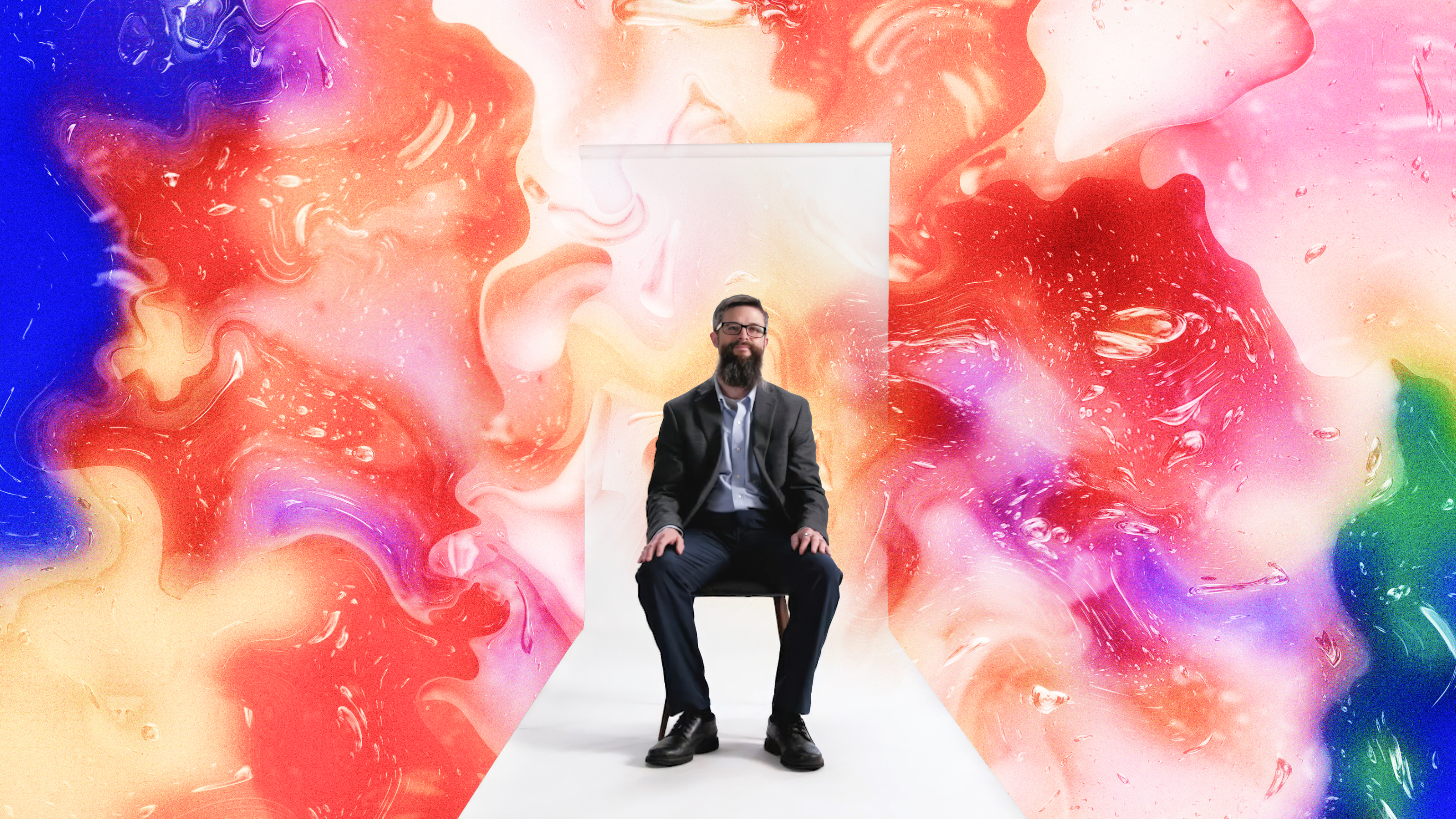 A man in a suit sits on a chair in front of a white door, surrounded by a vibrant, abstract swirl of red, pink, blue, yellow, and green colors.