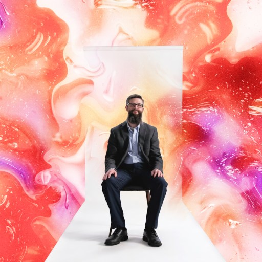 A man in a suit sits on a chair in front of a white door, surrounded by a vibrant, abstract swirl of red, pink, blue, yellow, and green colors.