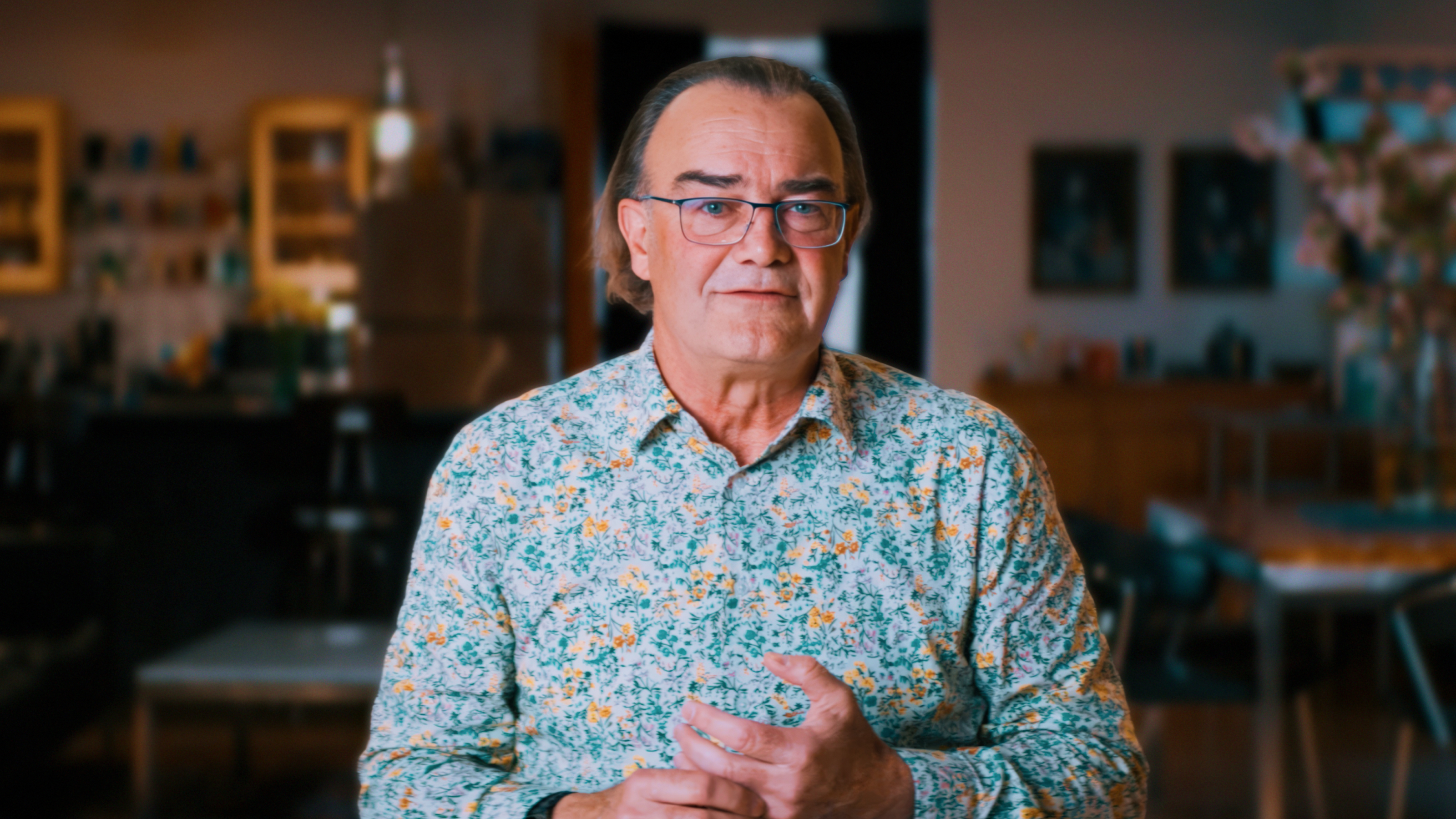 A middle-aged man with glasses and long hair, wearing a floral shirt, stands indoors in a warmly lit room with blurred background furniture.