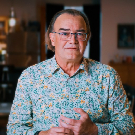 A middle-aged man with glasses and long hair, wearing a floral shirt, stands indoors in a warmly lit room with blurred background furniture.