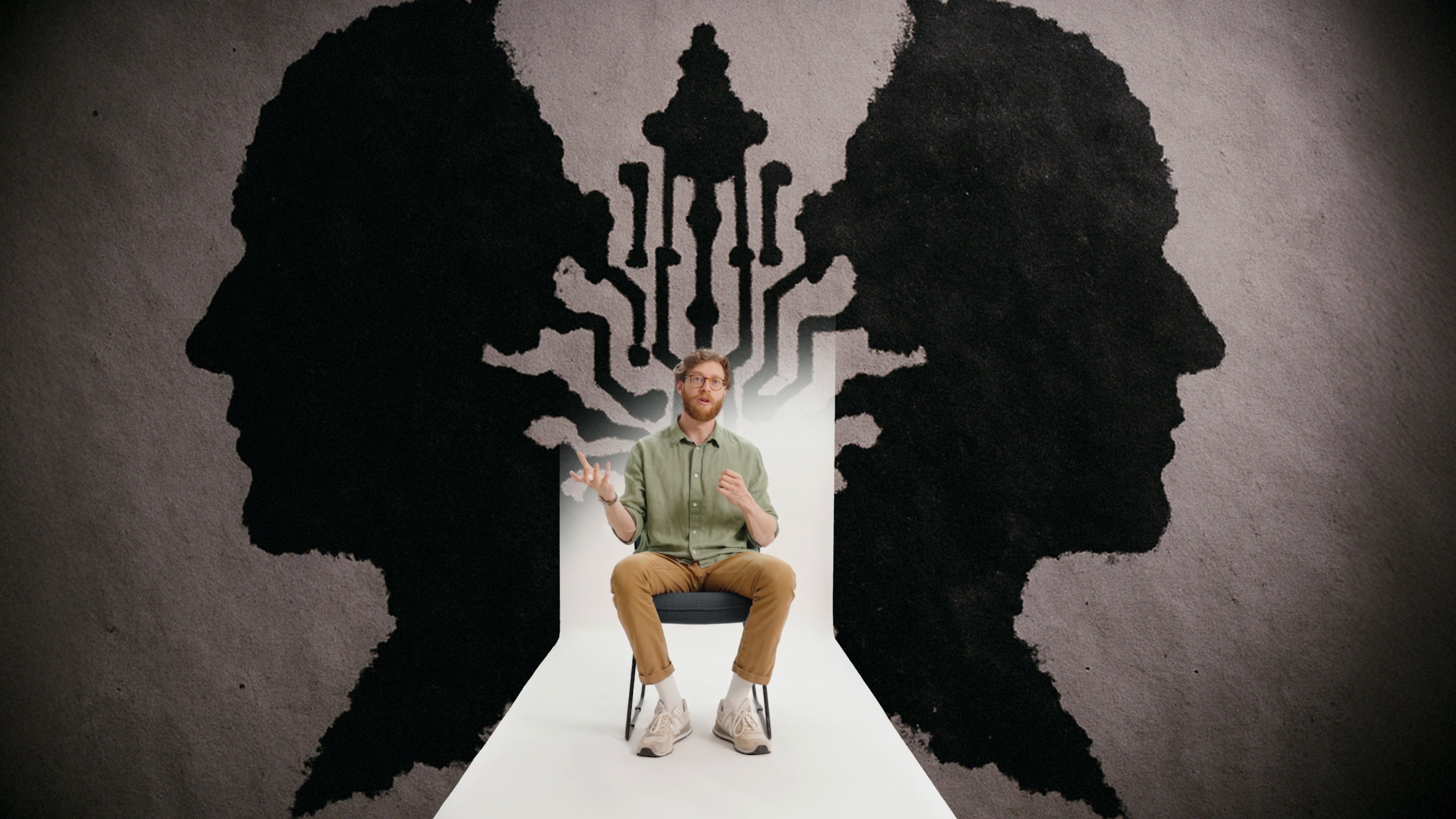 A man sits on a chair in front of a wall featuring abstract black silhouettes of two opposing heads and interconnected lines between them.