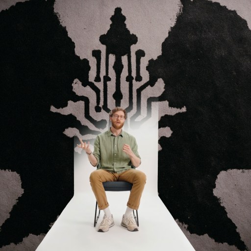 A man sits on a chair in front of a wall featuring abstract black silhouettes of two opposing heads and interconnected lines between them.