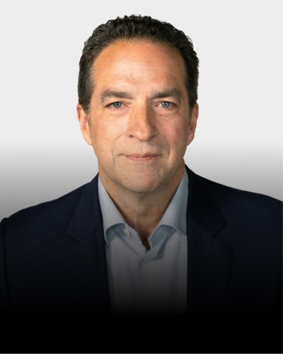 A man with short dark hair wearing a navy blazer and light blue shirt facing the camera against a plain light background.