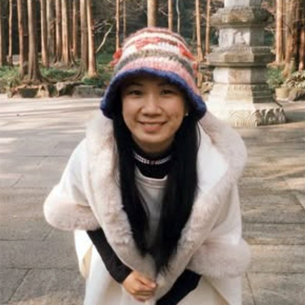 A woman in a white coat and colorful knit hat smiles outdoors in a wooded area with stone structures in the background.