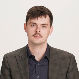 A man with short dark hair and a mustache wears a dark checked blazer over a navy shirt, posing against a plain light background.