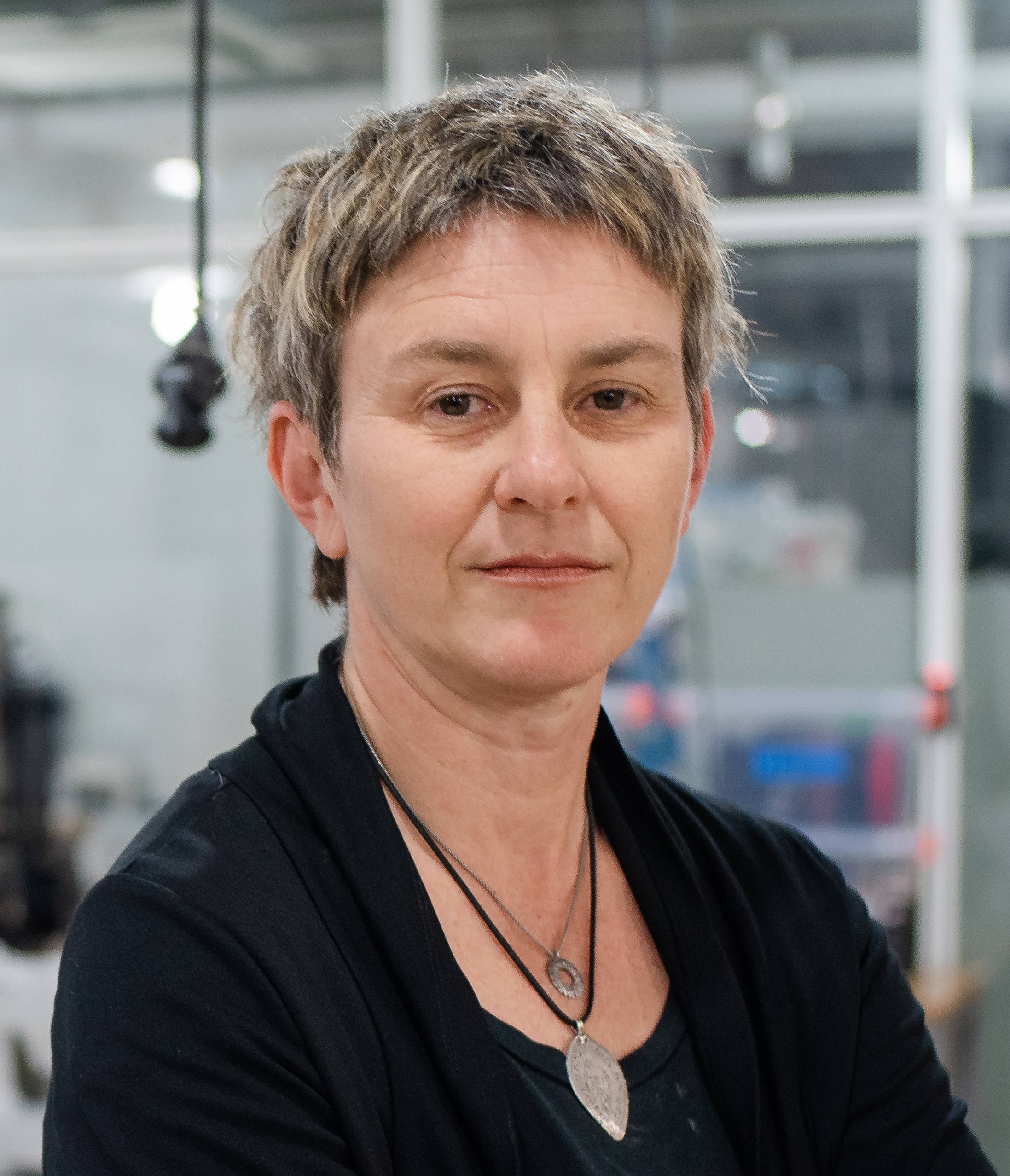 A person with short, textured hair and a neutral expression stands indoors, wearing a black top and two necklaces. The background shows a modern, glass-walled workspace.